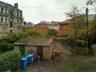 Back court behind Glasgow tenements, with communal bin store, drying poles and the rear gable of a sandstone tenement showing cast-iron downpipes